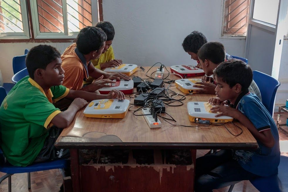 A group of six students are seated in two rows of three on either side of a wooden table. On the table are six Annies, one for each student. There is wiring on the table to connect the Annies to the power outlets