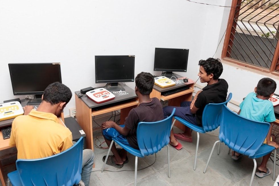 Three children sit in a classroom. They are at desks with computer monitors in front of the, beside which there are Annies. To the right of the image, there is another child sitting in front of his Annie, just below a window