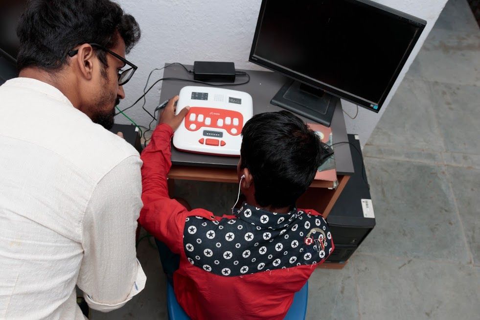 A man in a white shirt and glasses leans forward to talk to a student in a red patterned shirt. The student is seated at a table with a computer and an Annie in front of him 