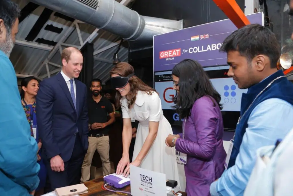 Duchess Catherine Middleton is blindfolded as she uses Annie, the Braille self-learning innovation. Prince William looks on, smiling. They are both formally dressed. Sanskriti Dawle and Aman Srivastava, co-founders of Thinkerbell Labs, showcase the device as others watch.