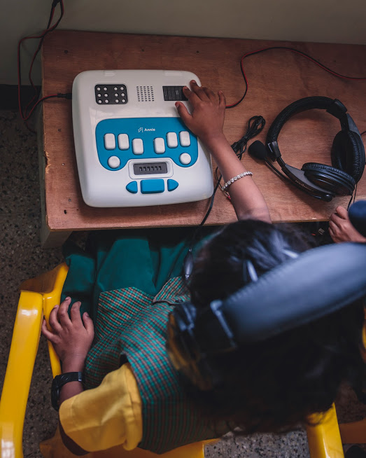 A visually impaired child is in her uniform, seated with an Annie in front of her. The child has headphones on