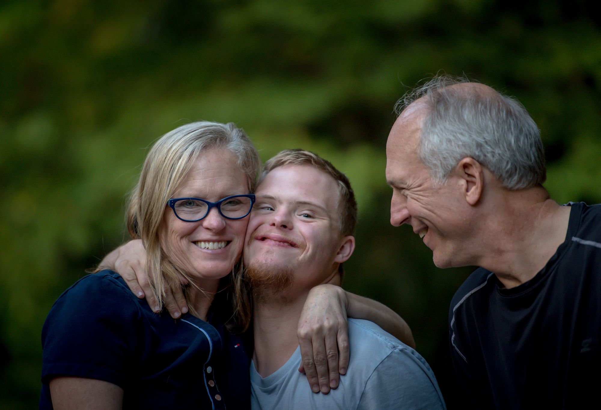 A smiling woman has her arm on a smiling young man's shoulder, her son. The man's father looks on in profile, smiling