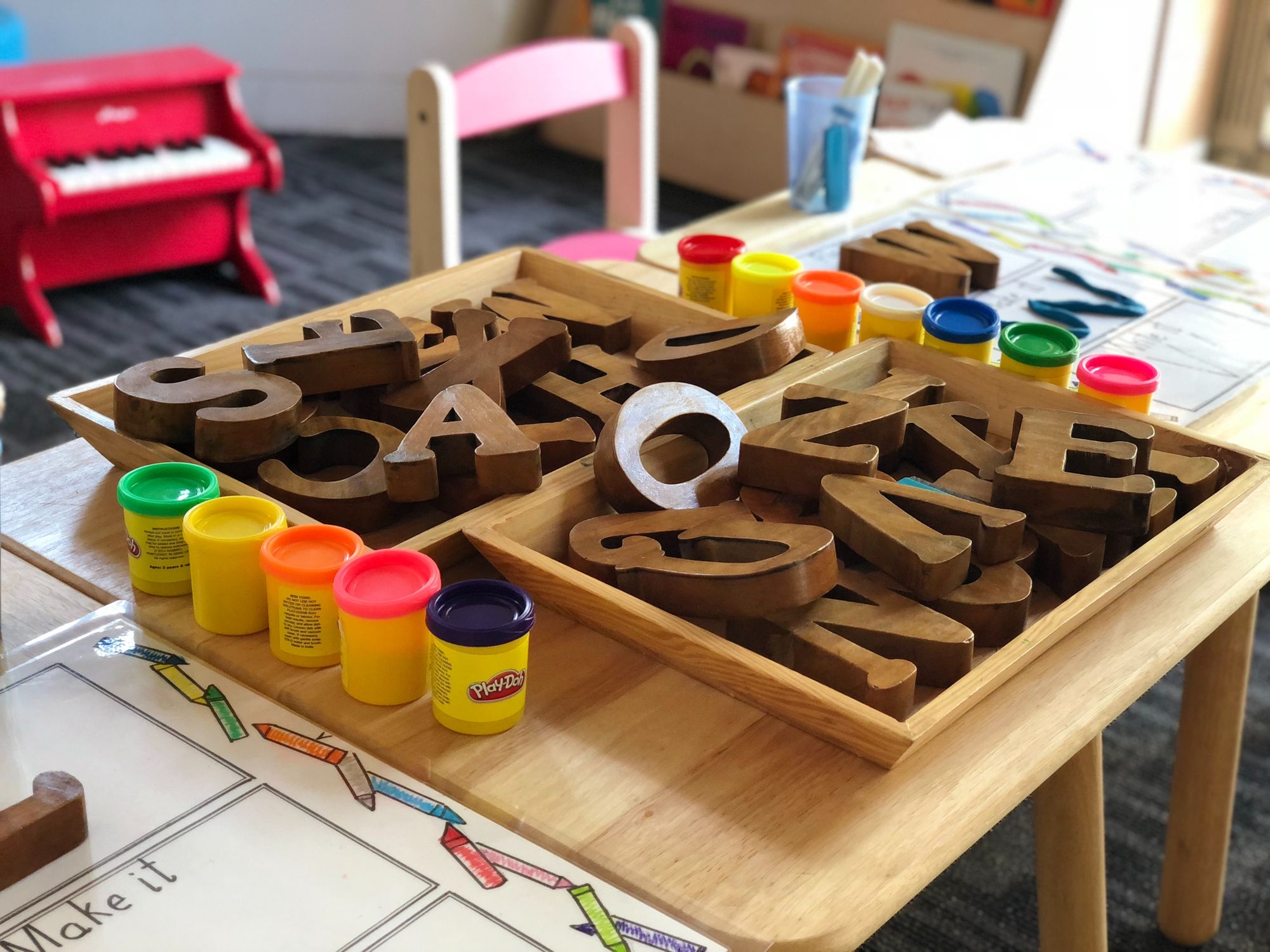 Wooden blocks of the English alphabet are placed in two trays on a table. There are canisters of Play Doh on both sides of the trays
