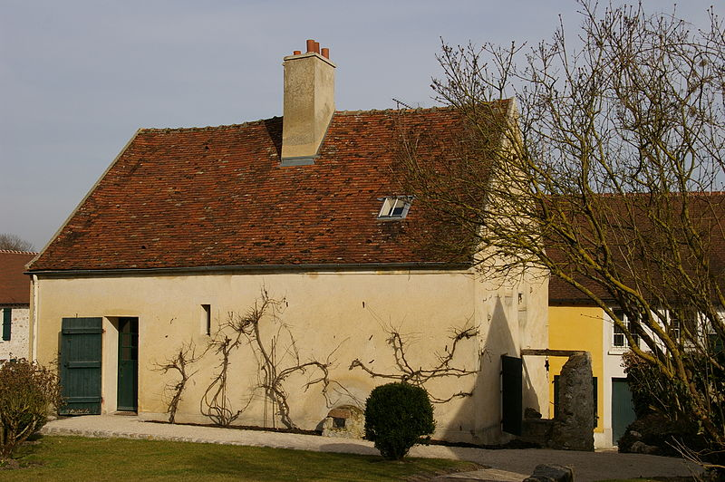 A single story house is seen. Its walls are beige, with red-tiled roof and a chimney. There is a manicured lawn and trees around the house