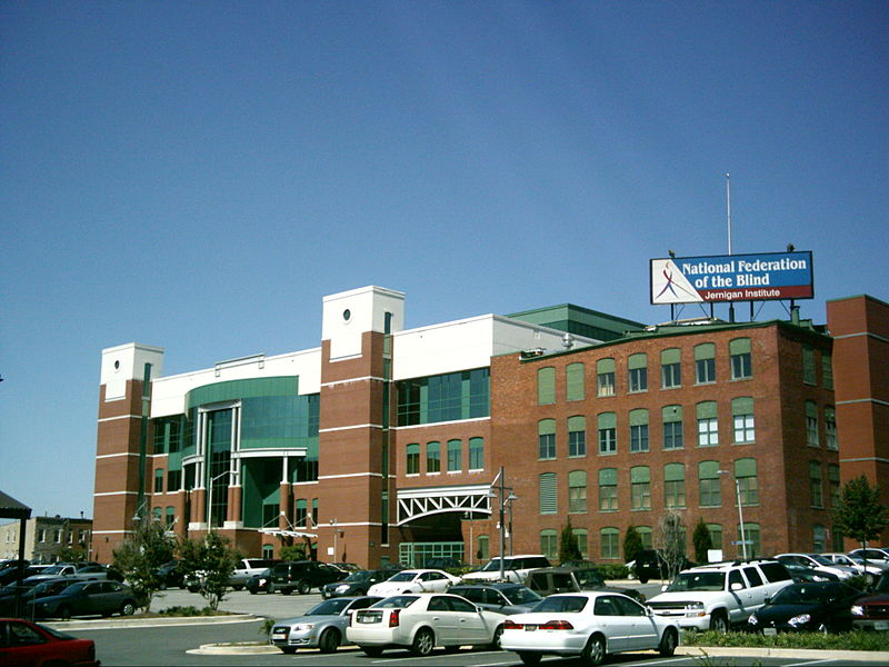 The building has a white terrace, and a brown brick and glass facade. A parking area with several vehicles is seen in front of the building