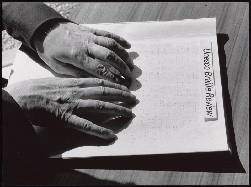  A close up of a man's hands while he's reading a text in braille. The paper shows, on top, the title "Unesco Braille Review" in bold type. 