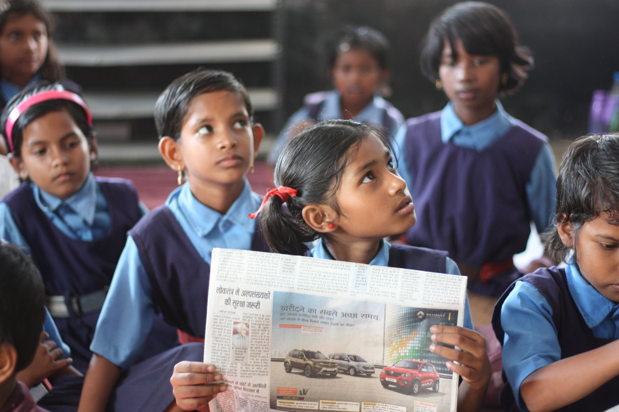 Curious Indian village girl students, sitting in the government school classroom, wearing a blue school dress and red ribbon.