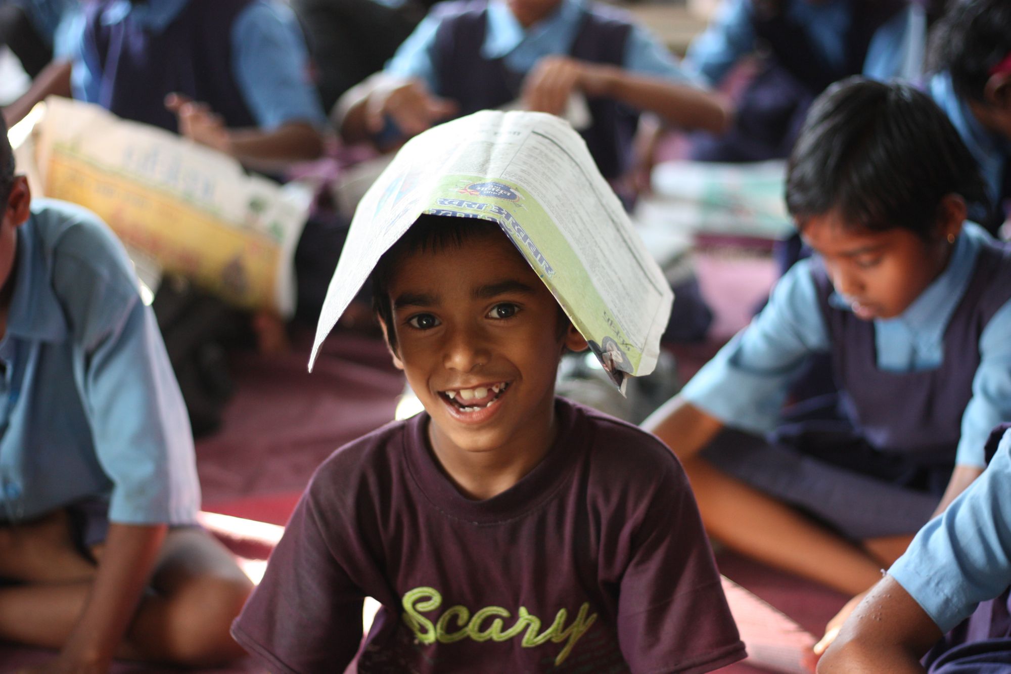 A boy is grinning at the camera, surrounded by other children in uniform in the background, with a book whose contents are unclear plopped on his head. 