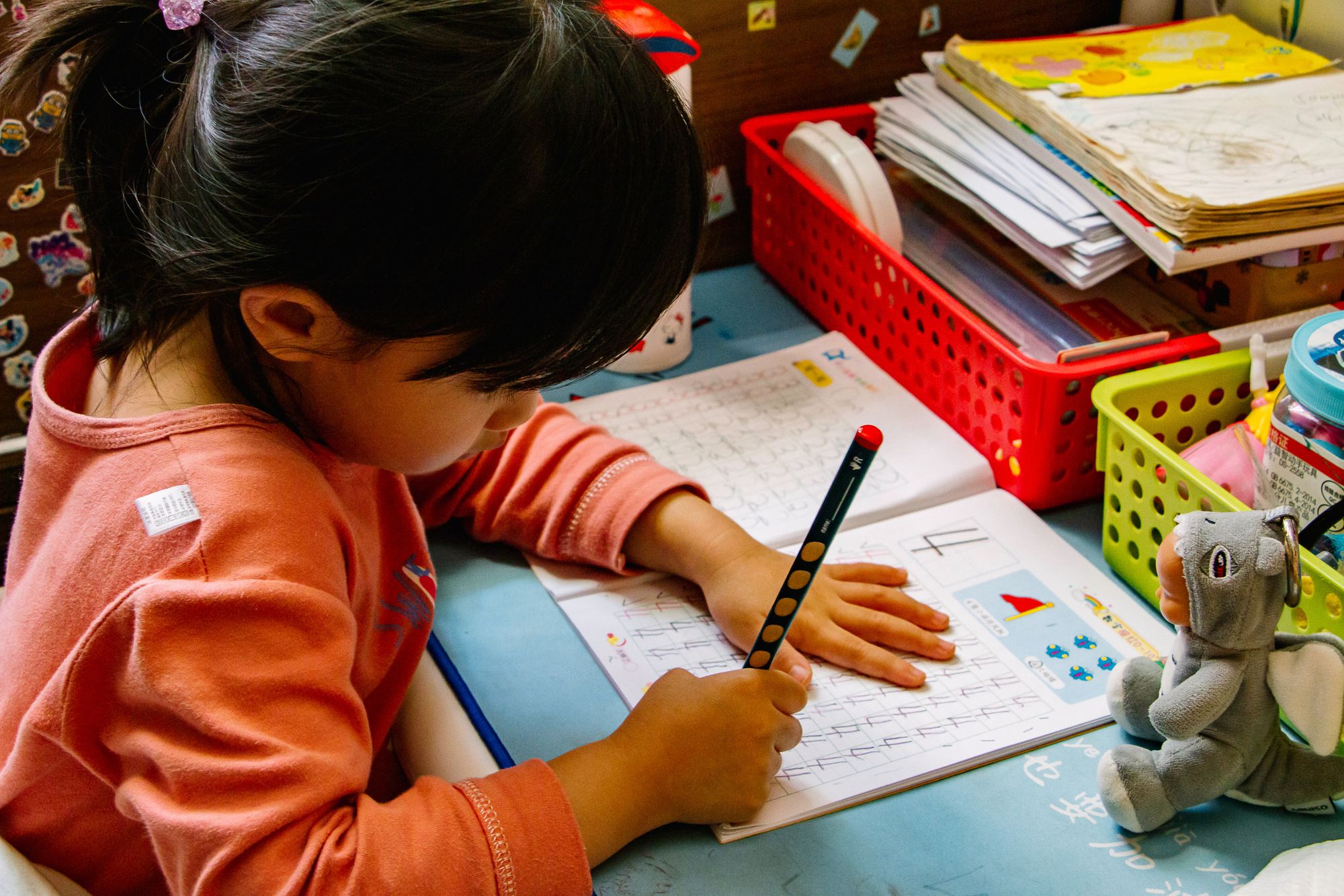 A girl in a pink t-shirt is practicing mathematics in her exercise book with a pencil. The table at which she is working is surrounded by trays with books as well as a soft toy 