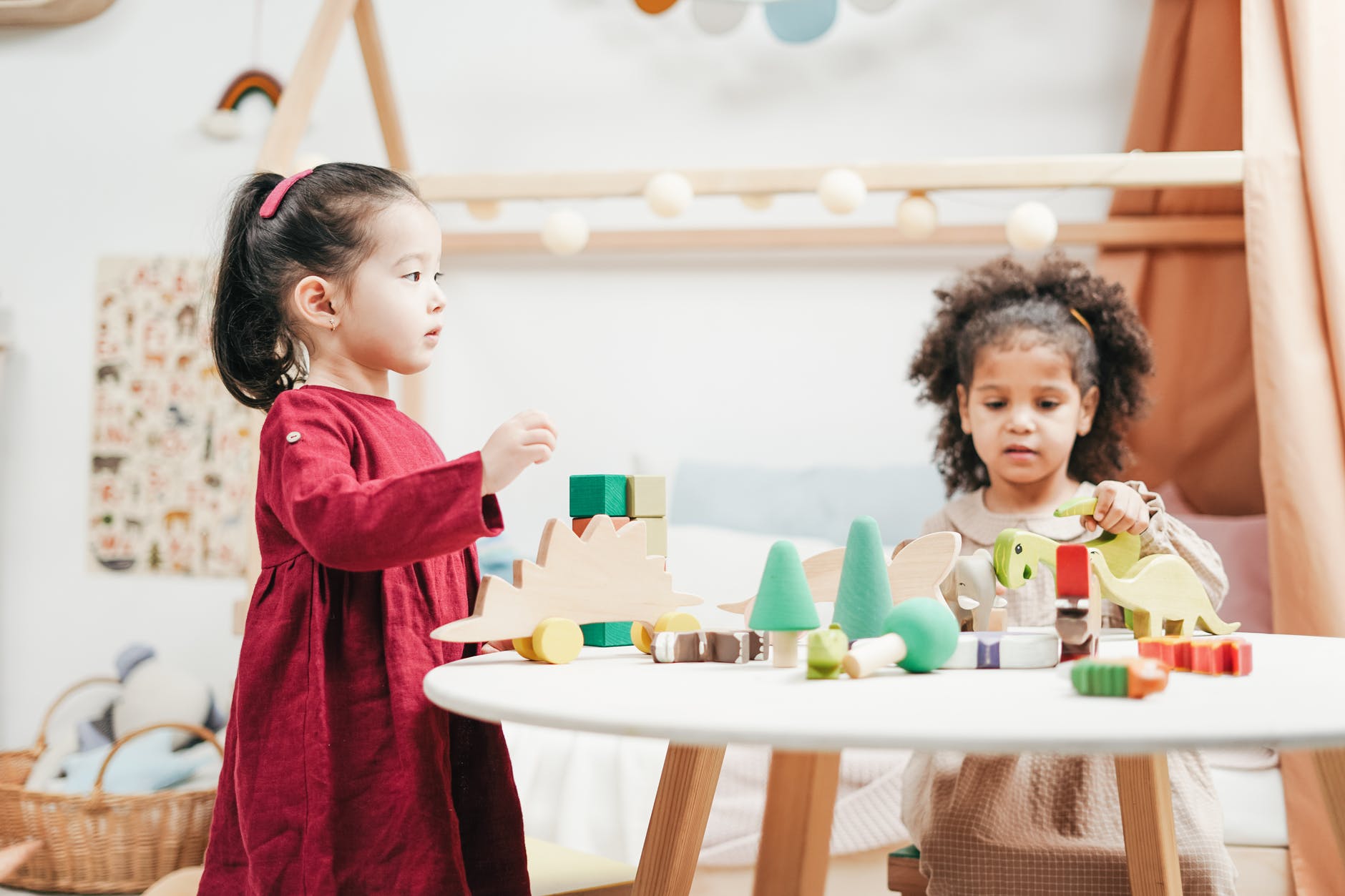 Two girls are playing with their toys. placed on a table. The toys include blocks of various colours, such as cyan, blue, and white, cones, as well as dinosaur figurines. 