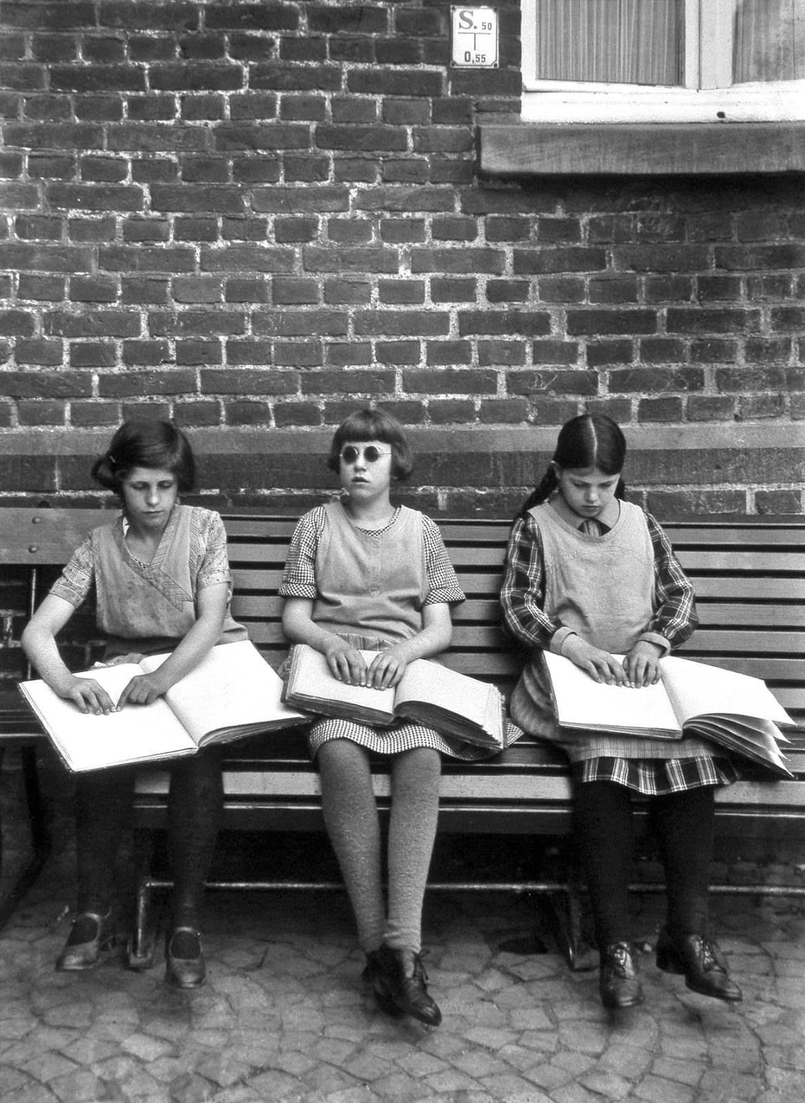 Three girls are sitting on a bench, reading from their Braille books. The image is in black and white