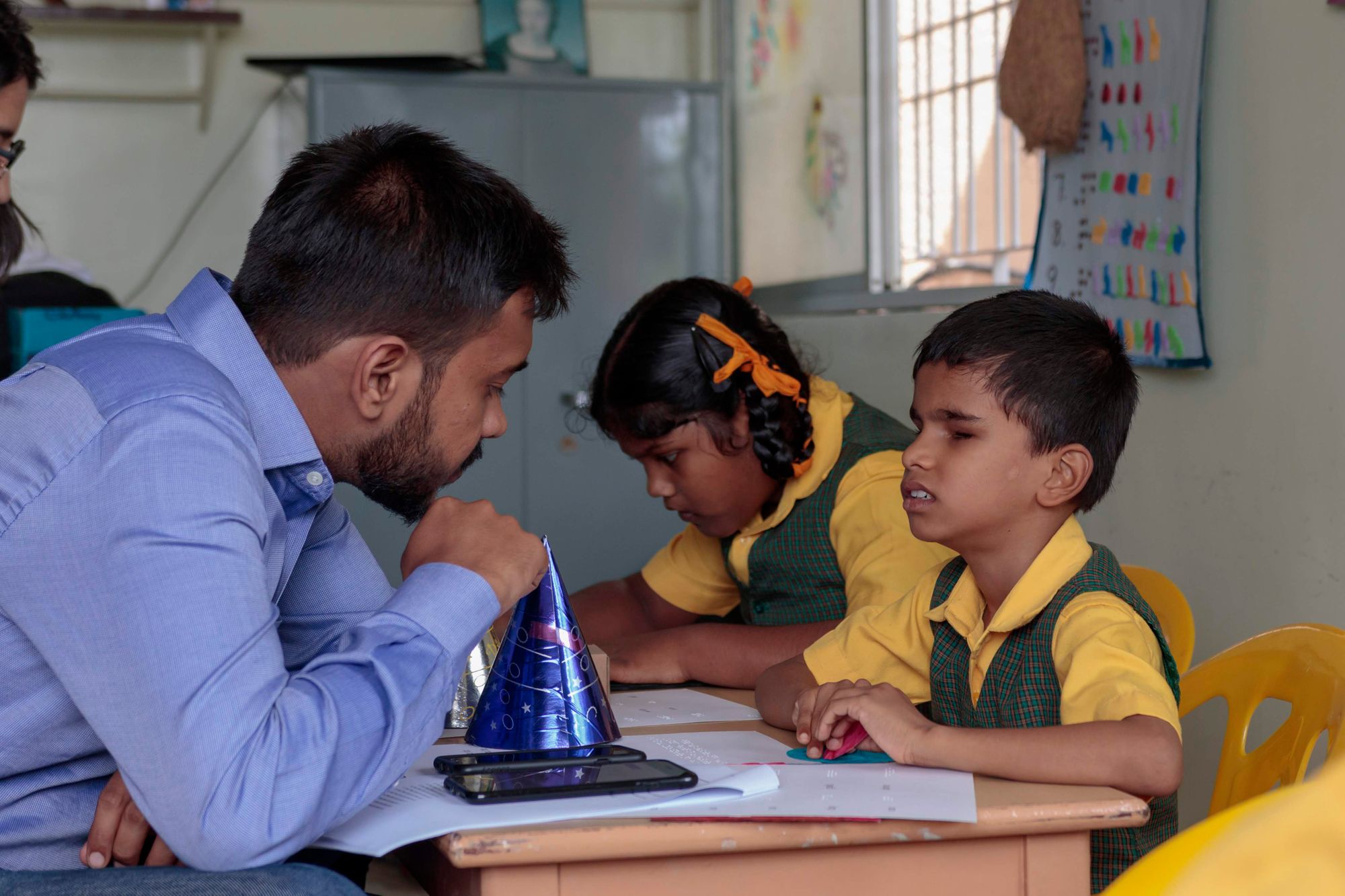 Two children are at their classroom tables reading Braille, while a man looks on