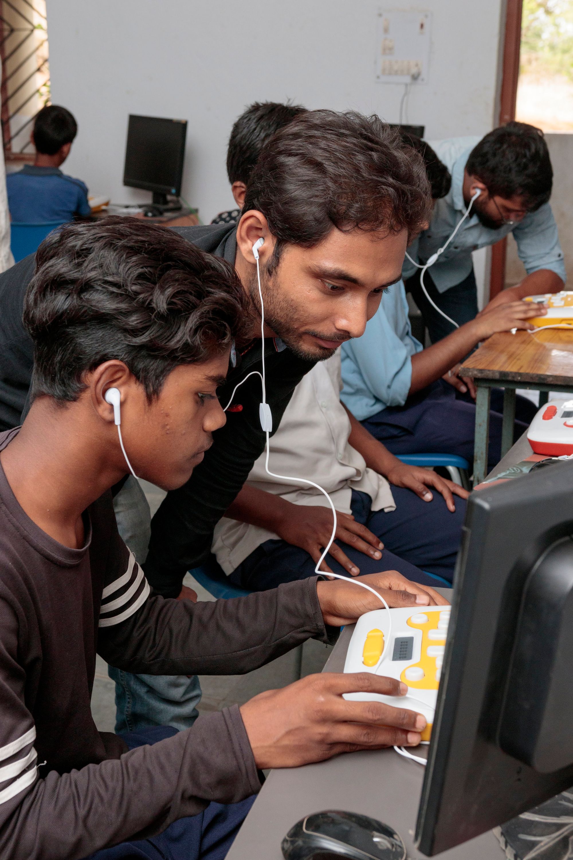 A teacher assists a student with using their Annie to learn Braille. Both of them have earphones plugged in