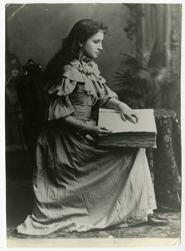 Portrait of Helen Keller as an adolescent reading a large braille book that is resting in her lap.