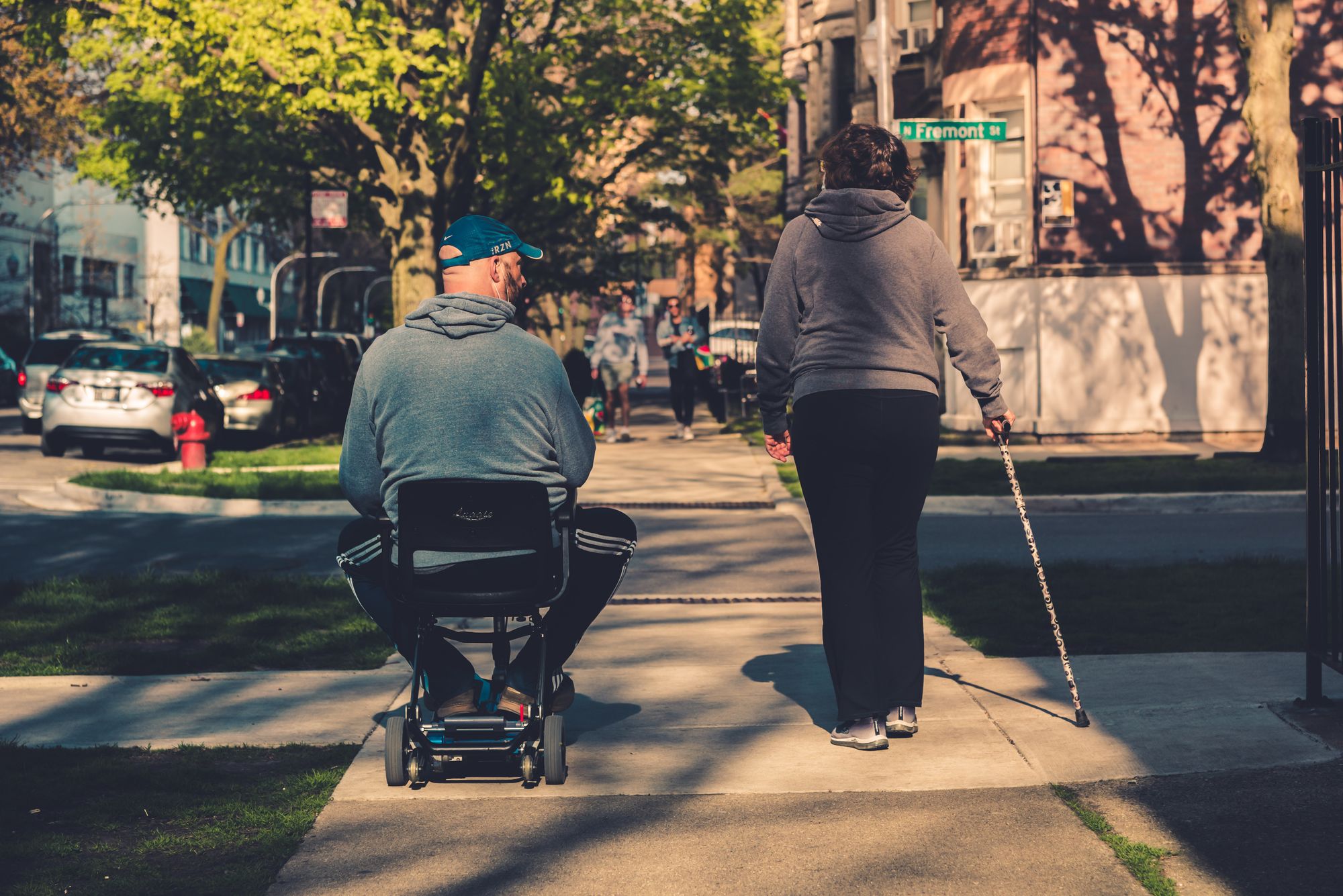 A man is riding in a wheelchair alongside a woman using a cane on a sidewalk. There are trees around, and parked cars. Two blurred figures are seen walking towards the camera.